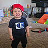 Jace is registered to the contest to win money with this photo: toddler, child, boy, red_cap, black_pants, tshirt, barefoot, carpet, living_room, toys, television, lamp, smiling, standing, indoors, furniture, play_area, cushion, air_conditioner, happy