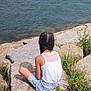 child, braided_hair, white_tank_top, denim_shorts, rocks, grass, water, boats, sea, sky, nature, coastline, outdoor, summer, relaxing, back_view, peaceful, shore, vacation, casual