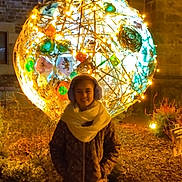 Eléonore participe au concours pour gagner de l'argent avec cette photo : art_installation, brick_wall, child, coat, decorative_lights, earmuffs, festive, glowing, ground, happy, lighting, night, outdoor, person, plants, scarf, shrubbery, smiling, sphere, warm_clothing
