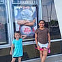 child, children, girl, smiling, posing, outdoor, movie_poster, building, window, concrete, sidewalk, doll, casual_clothing, sneakers, reflection, daylight, happy, toy, advertisement, fun