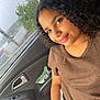 child, girl, car_interior, window, rain, raindrops, brown_clothing, bracelet, curly_hair, smile, portrait, seat, door_handle, car_door, natural_light, casual_wear, person, young, indoors, relaxed