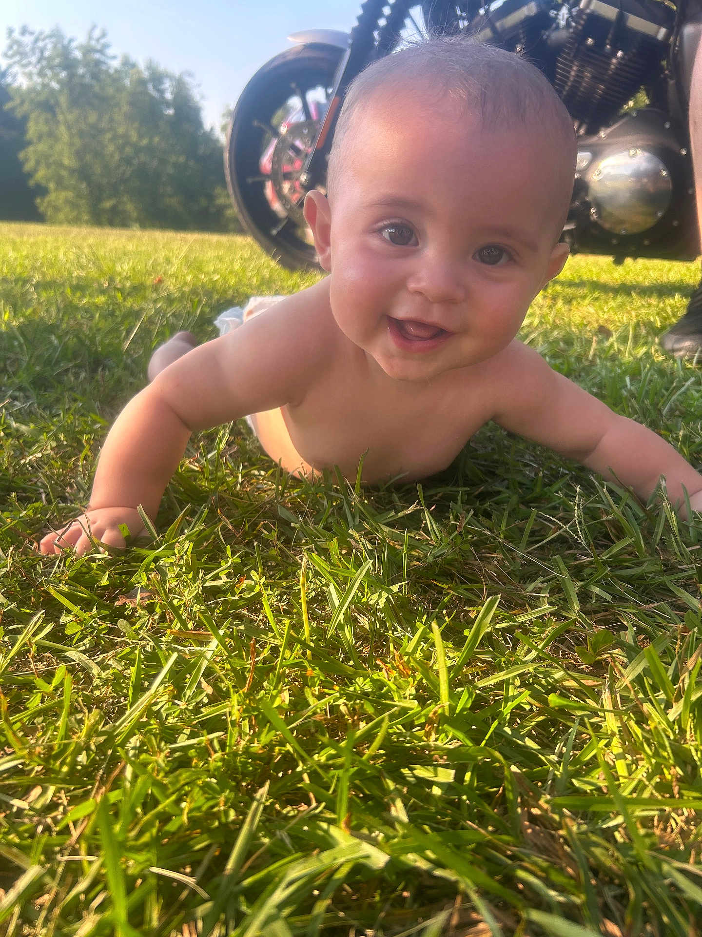 Noah joined the competition — help win amazing prizes! baby, child, grass, outdoor, sunlight, motorcycle, wheel, engine, smiling, crawling, happy, nature, greenery, skin, head, eyes, cheeks, person, daylight, background