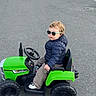 casual_clothing, child, cool, daytime, fun, jacket, outdoor, parking_lot, pavement, person, play, seat, sun_protection, sunglasses, toddler, toy_tractor, transportation, vehicle, wheel, young_child