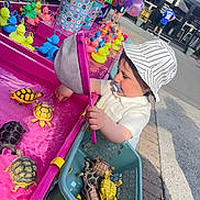 Victor a rejoint le concours — aidez-le/la à gagner de superbes lots ! toddler, child, toy_turtles, plastic_toys, fishing_game, net, basket, pacifier, hat, striped_hat, outdoor, carnival, balloons, rubber_ducks, pink, water, play, curious, person, sunlight