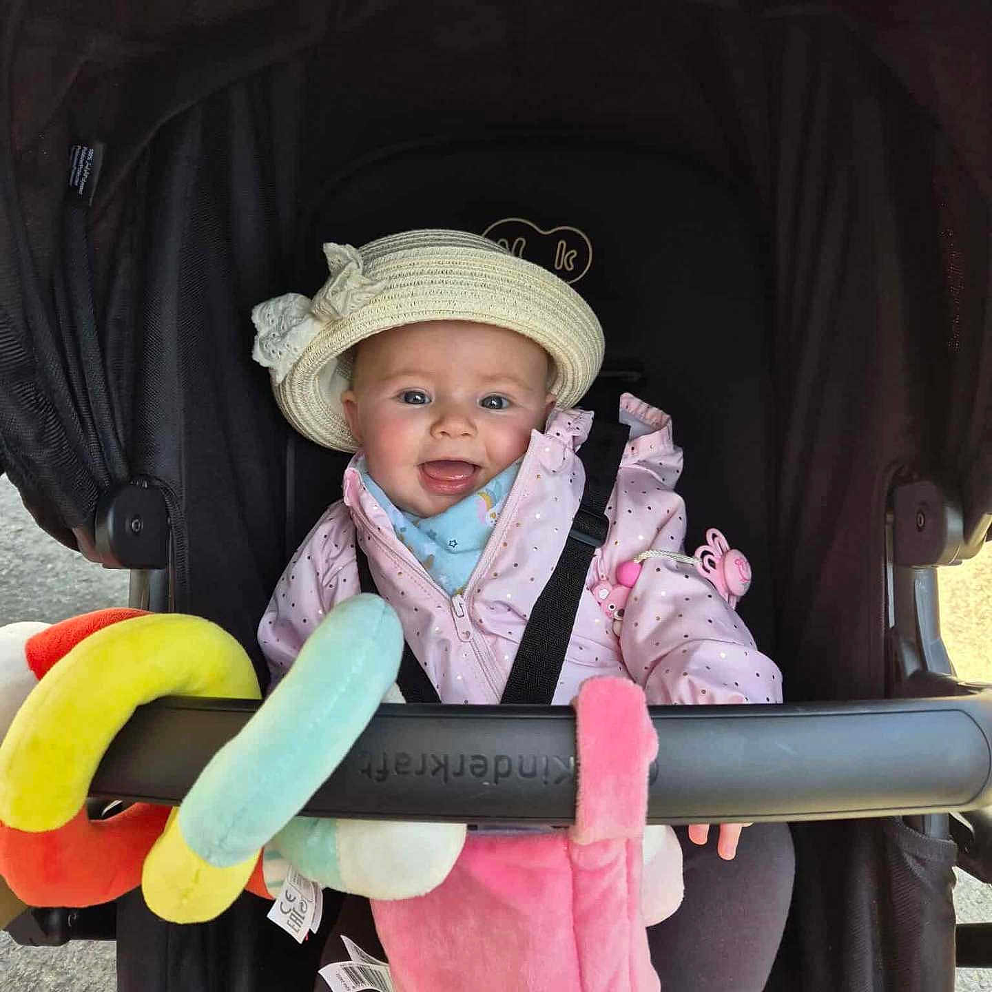 Leïla participe au concours pour gagner de l'argent avec cette photo : baby, baby_accessory, child, clothing, cute, face, happy, hat, headwear, infant, outdoor, person, pink_jacket, playful, plush, seatbelt, smile, stroller, sunlight, toy