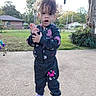 backyard, butterfly_pattern, casual_wear, child, concrete, curly_hair, daylight, expression, fence, grass, house, jacket, outdoor, pants, person, playful_pose, shoes, toddler, trees, young