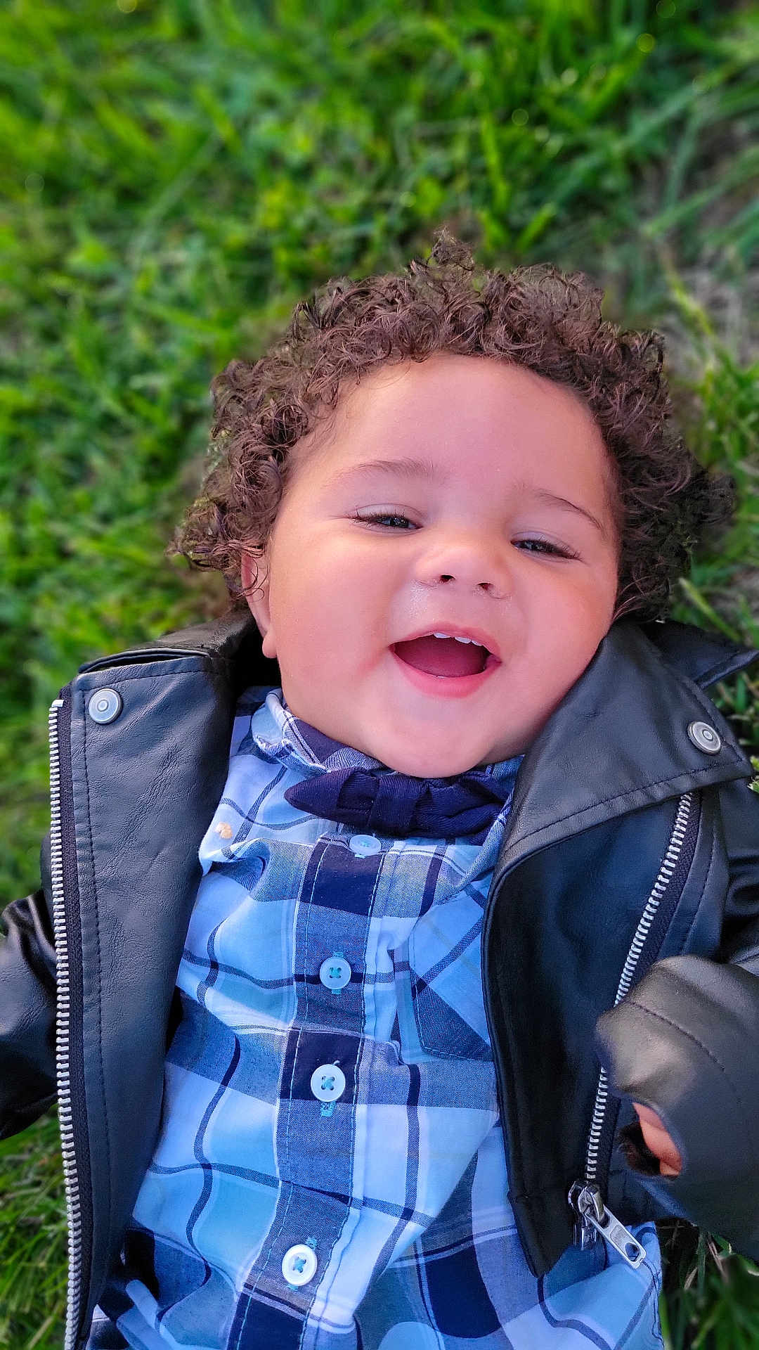 Tobiaz is registered to the contest to win money with this photo: child, toddler, smiling, curly_hair, plaid_shirt, leather_jacket, grass, outdoor, happy, portrait, baby, casual_clothing, cute, young, person, face, nature, playful, joyful, lying_down