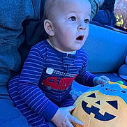 Hayson is registered to the contest to win money with this photo: baby, child, pajamas, striped_clothing, fire_truck_design, plush_toy, pumpkin, jack_o_lantern, couch, indoor, blue_lighting, person, hand, face, expression, seated, adult, blanket, cozy, soft_toy