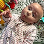 baby, child, onesie, striped_clothing, cartoon_print, blanket, toy, brush, hand, face, expression, lying_down, cute, infant, pattern, colorful, soft_toys, playtime, indoors, portrait
