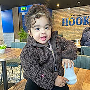 Liyana is registered to the contest to win money with this photo: child, toddler, smiling, curly_hair, brown_jacket, black_pants, indoor, cafe, table, wooden_table, pepper_grinder, plant, chair, brick_wall, signage, happy, person, seated, cozy, lighting