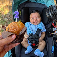 Rylan is registered to the contest to win money with this photo: baby, smile, muffin, candle, stroller, blue_clothing, baby_socks, hand, outdoor, celebration, birthday, food, face, seatbelt, car_seat, person, happy, child, ground, grass