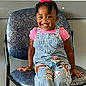 child, toddler, smiling, denim_overalls, pink_shirt, pink_bow, chair, indoor, happy, cute, sneakers, young_girl, seated, carpet, patterned_chair, joyful, portrait, casual_clothing, hair_braids, playful