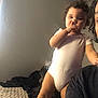 toddler, child, onesie, bed, indoor, person, curly_hair, skin, face, standing, picking_nose, adult_partial, expression, casual, clothing, light, shadow, wall, hair, room