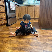 Scarlett is registered to the contest to win money with this photo: toddler, child, puppy, dog, kitchen, wooden_floor, cabinets, appliance, microwave, stove, curtains, window, barefoot, sitting, polka_dot, clothing, pet, indoor, floor, cute