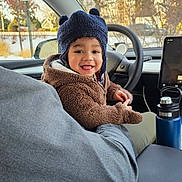 Edson is registered to the contest to win money with this photo: child, toddler, smiling, hat, winter_hat, fuzzy_jacket, car_interior, steering_wheel, water_bottle, seat, adult_arm, window, snowy_background, houses, dashboard, cozy, portrait, cute, candid, happy