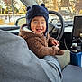 child, toddler, smiling, hat, winter_hat, fuzzy_jacket, car_interior, steering_wheel, water_bottle, seat, adult_arm, window, snowy_background, houses, dashboard, cozy, portrait, cute, candid, happy