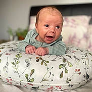 Jude is registered to the contest to win money with this photo: baby, infant, pillow, floral_pattern, bedroom, smiling, person, cute, happy, child, clothing, bed, head, face, hands, indoors, soft_focus, expression, lying_down, cozy