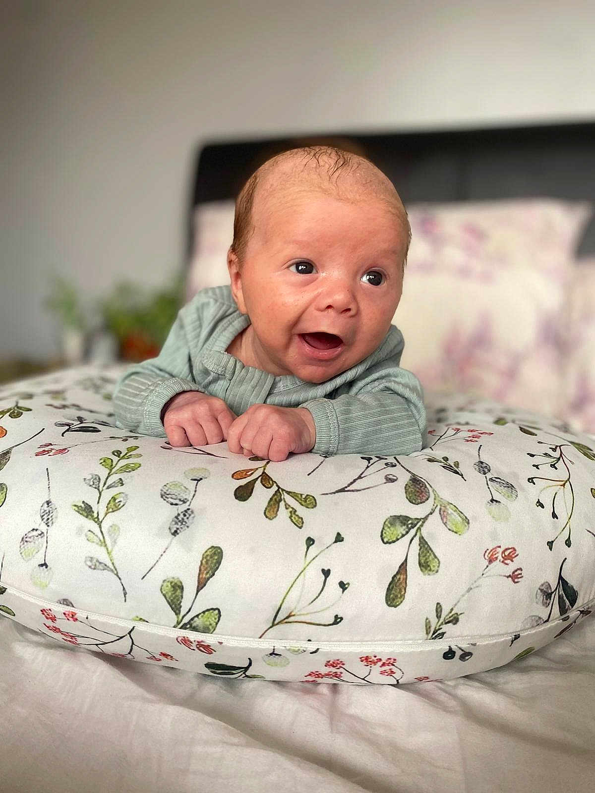 Jude is registered to the contest to win money with this photo: baby, infant, pillow, floral_pattern, bedroom, smiling, person, cute, happy, child, clothing, bed, head, face, hands, indoors, soft_focus, expression, lying_down, cozy