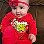baby, child, red_clothing, headband, bow, grinch, holiday_theme, pajamas, smiling, happy, couch, indoor, person, face, blue_eyes, cute, infant, sitting, cozy, playful