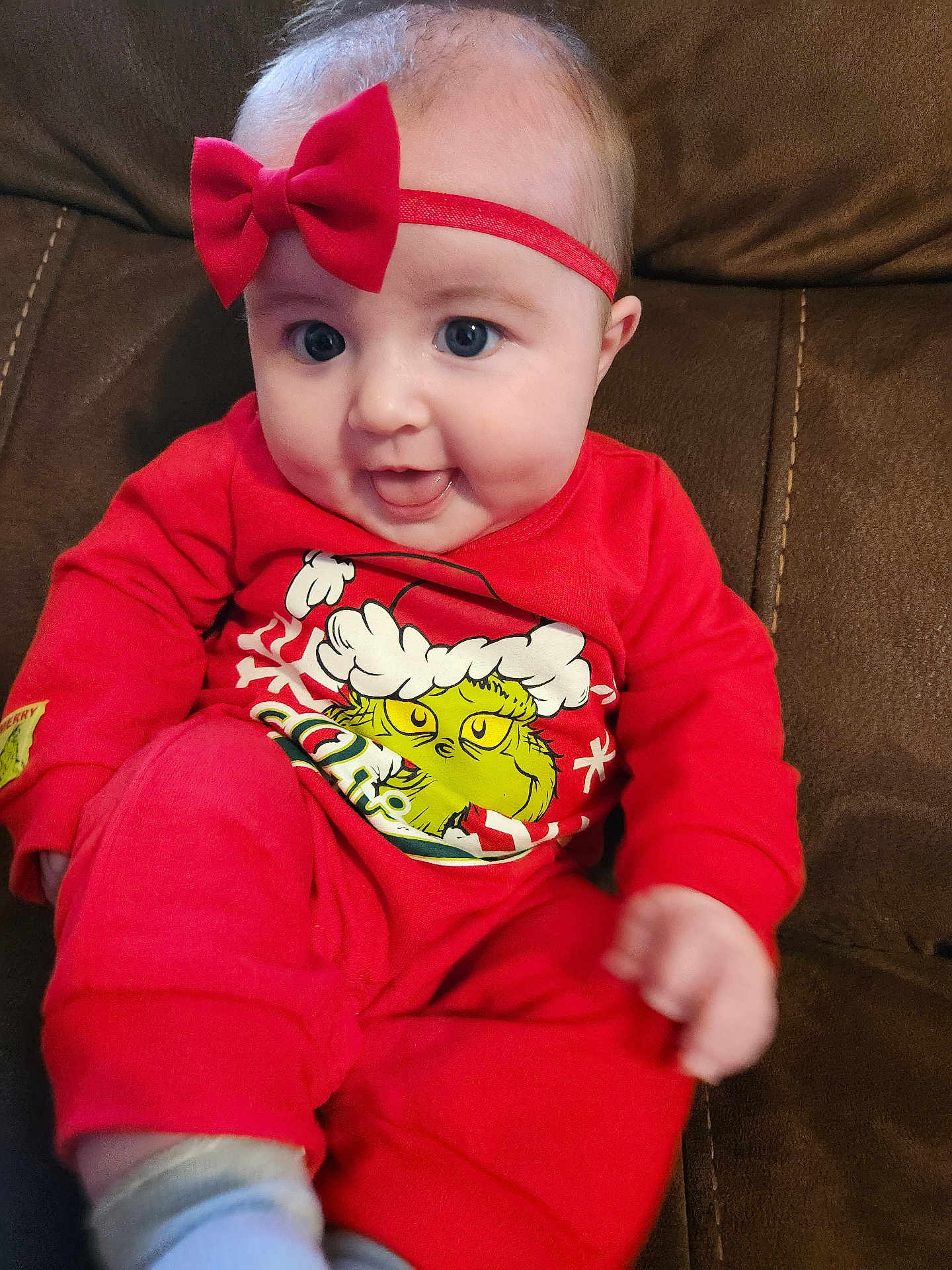 Emma is registered to the contest to win money with this photo: baby, child, red_clothing, headband, bow, grinch, holiday_theme, pajamas, smiling, happy, couch, indoor, person, face, blue_eyes, cute, infant, sitting, cozy, playful