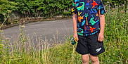Alistair is registered to the contest to win money with this photo: block, boy, brick_wall, child, daytime, dinosaur_pattern, grass, greenery, house, nature, outdoor, plants, residential_area, road, shoes, shorts, smile, standing, stone, tshirt