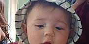 Stephen is registered to the contest to win money with this photo: baby, child, hoodie, striped_clothing, ears_on_hood, indoor, woman, person, face, closeup, portrait, seated, blond_hair, casual_clothing, expressionless, background_people, soft_light, home_interior, window, curtain