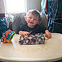 Stephen is registered to the contest to win money with this photo: baby, high_chair, toy_truck, gift_wrap, smiling, child, indoor, window, messy, table, food_crumbs, clothing, face, person, seat, happy, young_child, present, playing, home