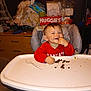 Stephen is registered to the contest to win money with this photo: toddler, high_chair, red_shirt, messy, snack, crumbs, plastic_spoon, happy, smiling, indoor, diapers, clutter, drawer, household_items, face, child, person, eating, seat, graco