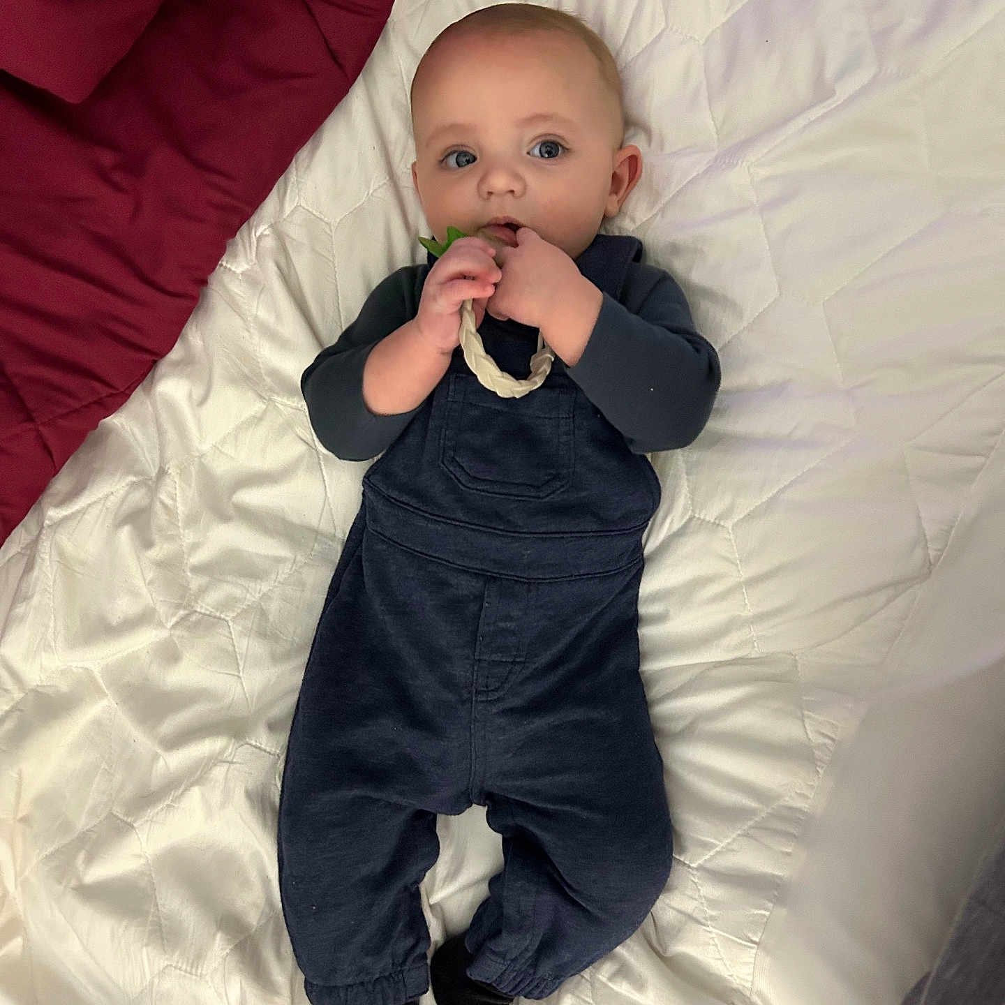 Solomon is registered to the contest to win money with this photo: baby, bed, black_socks, blanket, blue_clothing, blue_eyes, braided_ring, child, curious, cute, face, hand, indoor, infant, lying_down, overalls, quilted_bedspread, sleepwear, soft_lighting, toy