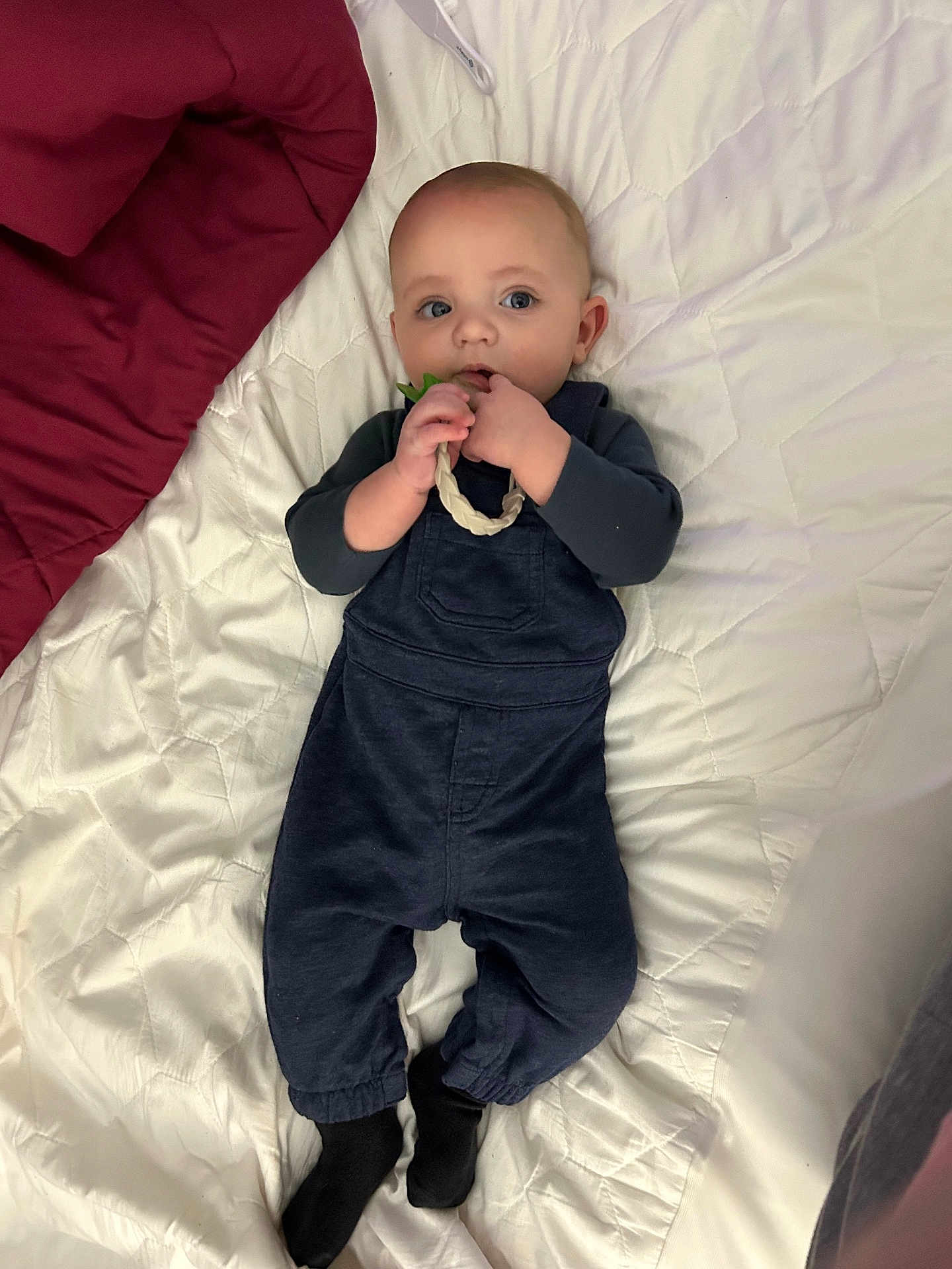 Solomon is registered to the contest to win money with this photo: baby, infant, child, overalls, blue_clothing, black_socks, bed, quilted_bedspread, toy, braided_ring, lying_down, cute, curious, hand, face, blue_eyes, indoor, soft_lighting, blanket, sleepwear