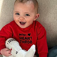 Solomon is registered to the contest to win money with this photo: baby, cheek, couch, cute, ear, eyes, fabric_texture, hand, happy, holding_foot, indoor, infant, leg, portrait, red_shirt, sitting, smiling, sock, text_on_clothing, toddler