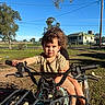 child, toddler, quad_bike, outdoor, sunny, grass, tree, sky, house, fence, handlebars, vehicle, person, playing, daylight, expression, clothing, summer, nature, park
