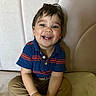 toddler, child, smile, happy, sitting, blue_shirt, striped_clothing, khaki_pants, indoor, couch, furniture, face, person, cute, young_child, hands, hair, portrait, casual, playful