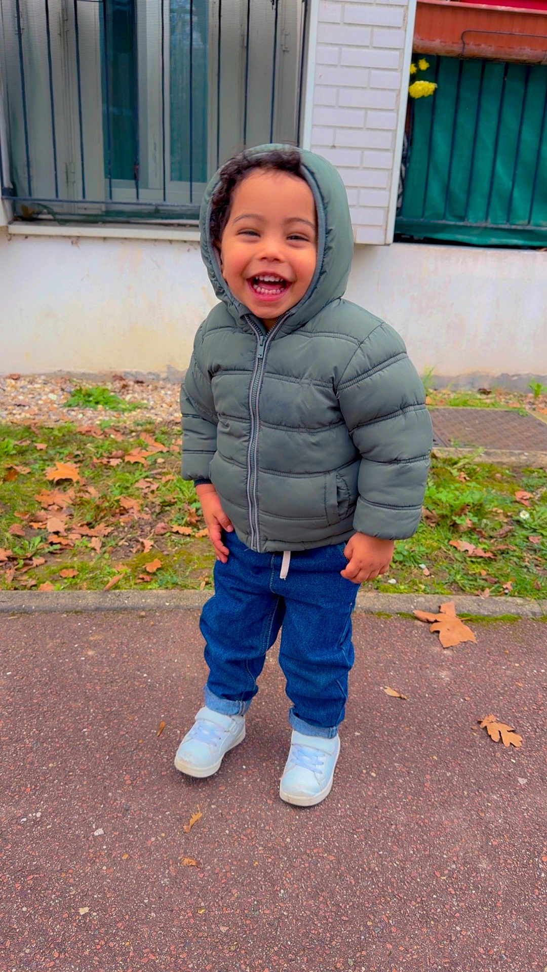 Adem a rejoint le concours — aidez-le/la à gagner de superbes lots ! toddler, child, smiling, hooded_jacket, blue_jeans, white_shoes, outdoor, pavement, grass, fallen_leaves, window, building, autumn, happy, young_child, casual_clothing, standing, daylight, curly_hair, person
