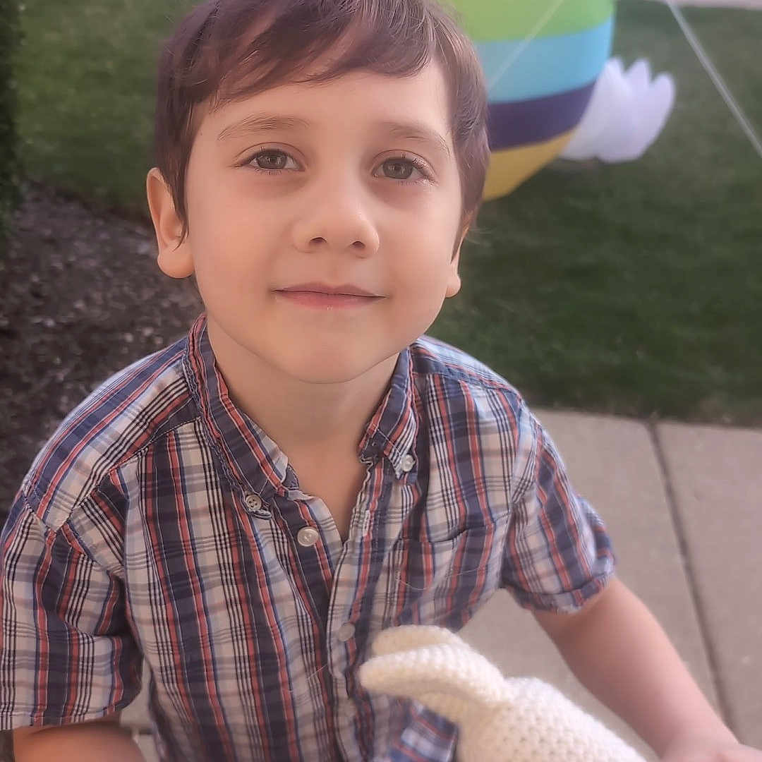 Ben is registered to the contest to win money with this photo: child, boy, scooter, balloon, bunny, toy, outdoor, sidewalk, grass, plaid_shirt, short_hair, smiling, daylight, playful, knitted_toy, colorful, person, casual_clothing, background, happy