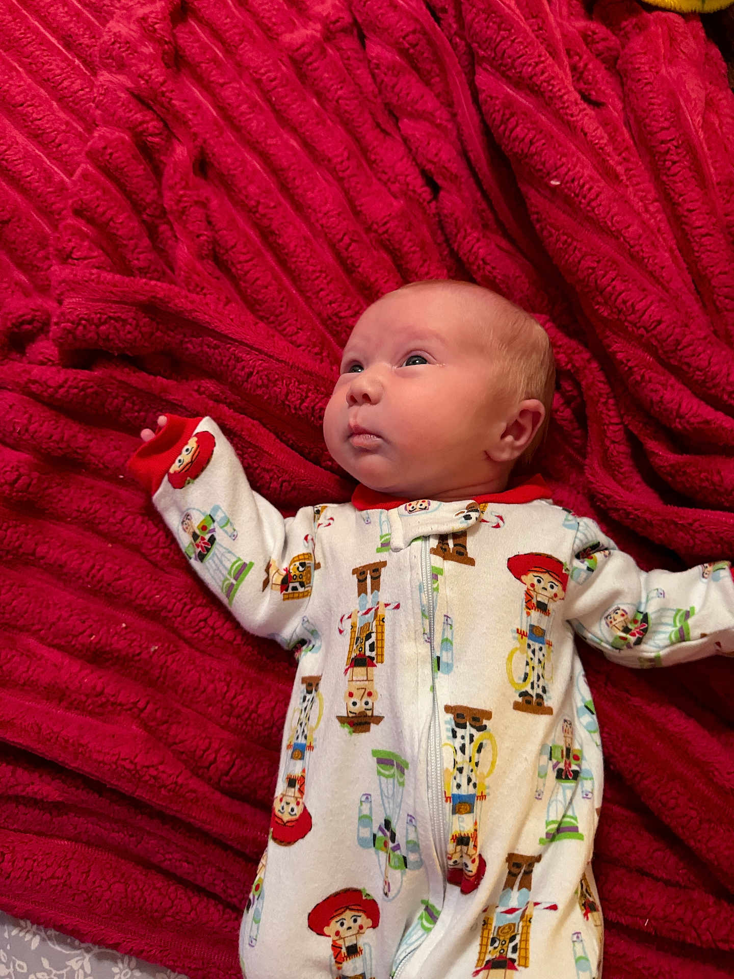 Juniper is registered to the contest to win money with this photo: baby, infant, blanket, red, sleepwear, toy_story, pajamas, face, expression, child, cute, lying_down, soft_texture, bed, patterned_clothing, newborn, person, warm, cozy, indoors