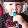 child, toddler, portrait, indoors, baseball_cap, cardinals_cap, lollipop, candy, blue_lollipop, serious_expression, blond_hair, blue_eyes, black_shirt, new_balance, hands, sitting, bedroom, furniture, close_up, candid_photo