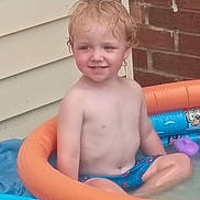 Tyler joined the competition — help win amazing prizes! child, toddler, smiling, portrait, outdoor, backyard, swimming_pool, inflatable_pool, water, summer, bare_chested, swim_shorts, seated, blond_hair, blue_pool, orange_float, cartoon_design, brick_wall, house_siding, plastic_tub