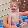 child, toddler, smiling, portrait, outdoor, backyard, swimming_pool, inflatable_pool, water, summer, bare_chested, swim_shorts, seated, blond_hair, blue_pool, orange_float, cartoon_design, brick_wall, house_siding, plastic_tub