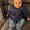 baby, child, puffer_jacket, jeans, socks, couch, sofa, indoor, curious, expression, seated, casual_clothing, brown, blue, cozy, infant, portrait, home, person, cute