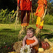 Amara joined the competition — help win amazing prizes! baby, pumpkin, flower, scarecrow, autumn, hay, grass, outdoor, playful, tongue_out, greenery, sunlight, fall_decor, child, nature, orange, yellow, brown, cute, seasonal