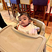 Aaira Ghosh is registered to the contest to win money with this photo: baby, bib, child, clothing, cute, expression, face, floor, furniture, headband, high_chair, indoor, infant, person, seat, stool, table, tile, wood, young