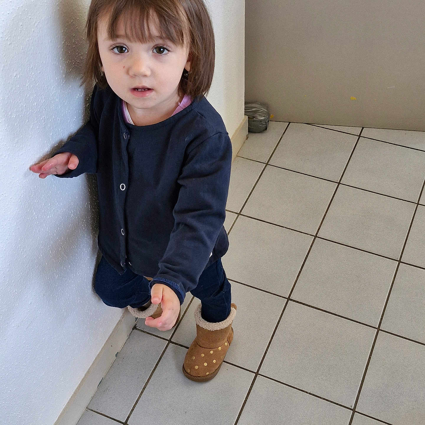 Julia a rejoint le concours — aidez-le/la à gagner de superbes lots ! boots, brown_boots, casual_clothing, child, curious_expression, floor_tiles, home_interior, indoor, light_skin, looking_at_camera, navy_cardigan, person, polka_dots, short_hair, small_child, standing, tile_floor, toddler, wall, young_child