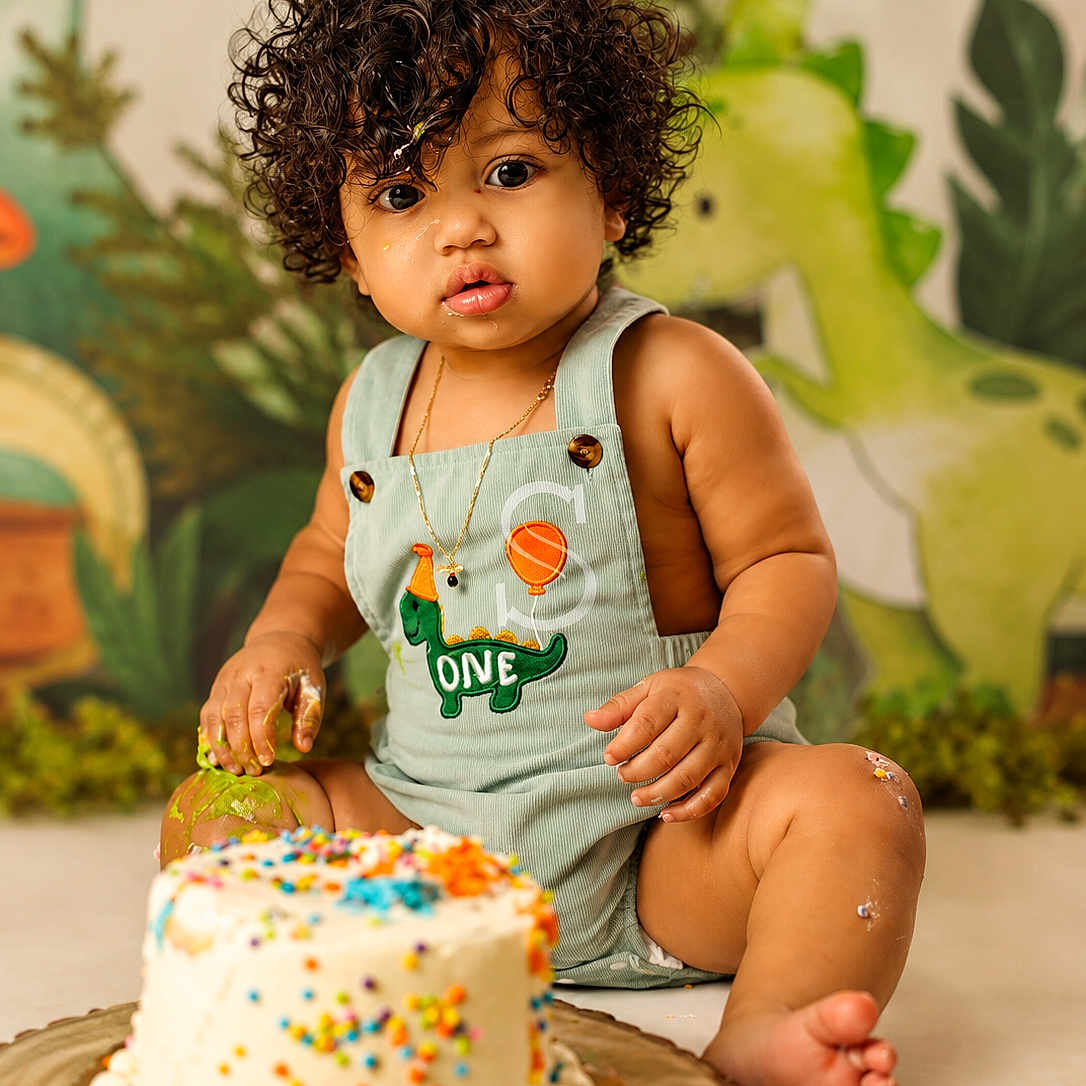 Eithan Milan joined the competition — help win amazing prizes! baby, birthday_cake, cake, celebration, child, colorful, curly_hair, cute, decor, dinosaur, food, green_romper, happy, indoors, party_hat, playful, portrait, sitting, sprinkles, toddler