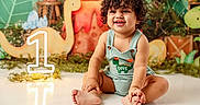 Eithan Milan is registered to the contest to win money with this photo: toddler, child, smiling, curly_hair, sitting, romper, dinosaur, birthday, number_one, decorations, greenery, playful, indoor, cute, happy, portrait, floor, celebration, baby, one_year_old