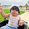 Eithan Milan joined the competition — help win amazing prizes! baby, casual_clothing, child, closeup, curly_hair, cute, daylight, denim_shorts, expression, gold_necklaces, grass, hand_gesture, outdoor, portrait, sidewalk, sitting, toddler, toy_car, white_cap, white_tank_top