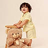 Eithan Milan is registered to the contest to win money with this photo: toddler, child, curly_hair, yellow_outfit, striped_clothing, standing, wooden_stool, teddy_bear, plush_toy, smiling, studio_background, indoor, cute, happy, playful, person, portrait, soft_lighting, childhood, innocence