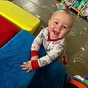 Barrett is registered to the contest to win money with this photo: baby, child, smiling, blue_cushion, yellow_cushion, red_cushion, playroom, toys, floor_mat, shelf, stuffed_toy, colorful, indoor, happy, pajamas, crawling, face, hands, wooden_floor, infant