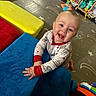 baby, child, smiling, blue_cushion, yellow_cushion, red_cushion, playroom, toys, floor_mat, shelf, stuffed_toy, colorful, indoor, happy, pajamas, crawling, face, hands, wooden_floor, infant