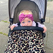 Mabel is registered to the contest to win money with this photo: baby, blue_eyes, headband, pink_headband, stroller, blanket, leopard_print, outdoor, sidewalk, infant, child, cute, person, young_child, hand, clothing, urban, seat, safety_bar, expression